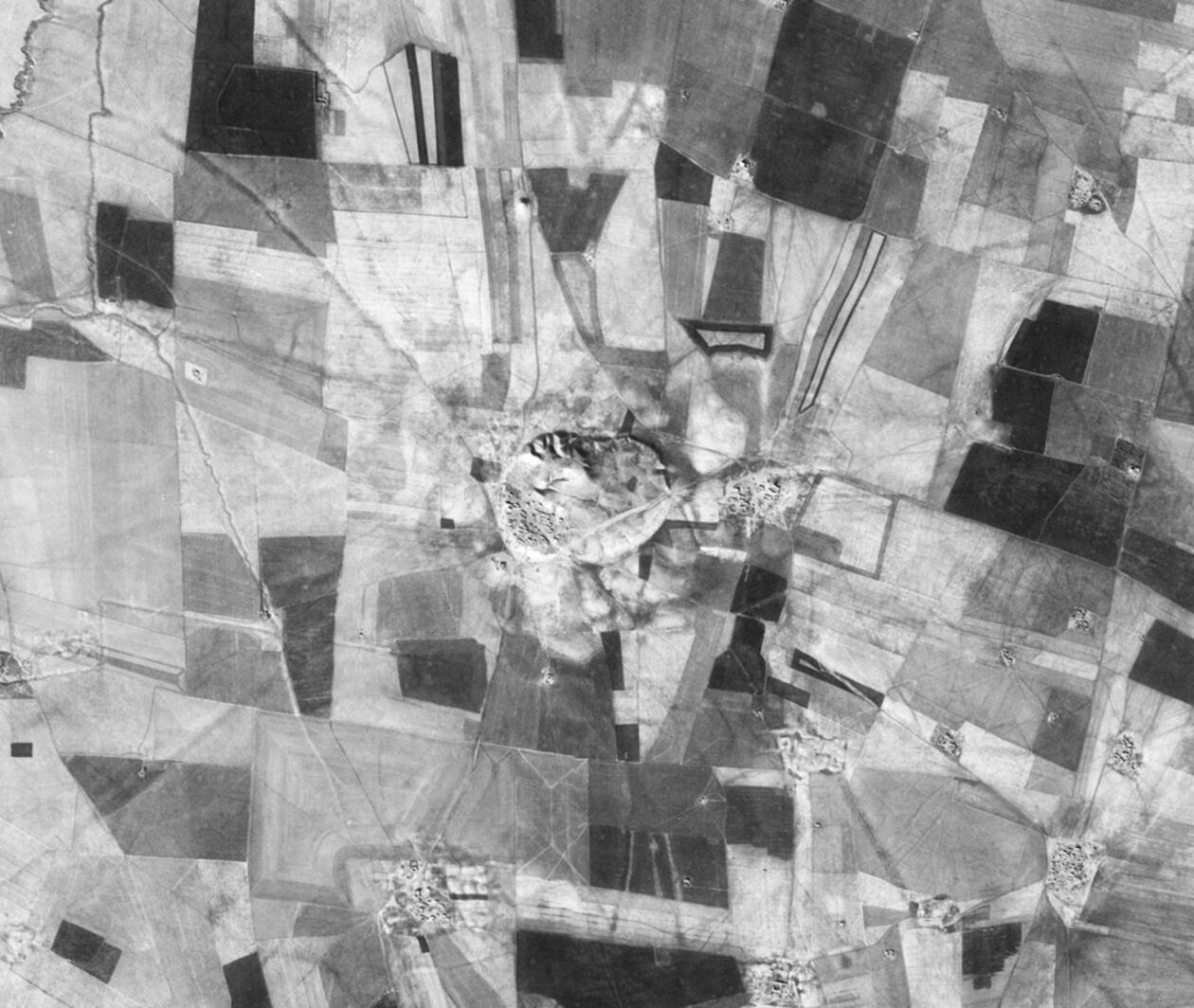 A black and white aerial photo of a large tell, surrounded by modern agricultural fields and small villages.