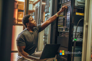 A male Azure data engineer sat in a sever room, connecting the laptop on his lap to the server tower next to him, to begin his data analytics. 