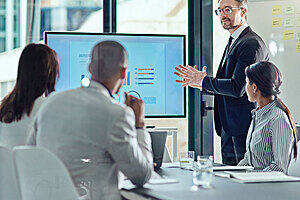 Three people sat at a table looking at a man standing in front of a screen with charts on it.