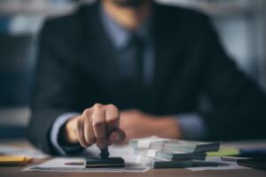 A person in a suit sitting at a desk and holding a stamp on one page of a financial contract.