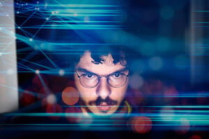 A young male computer programmer looking at his computer with overlay abstract finance economic graphs in the foreground.