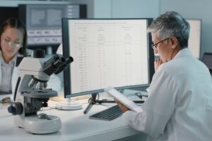 Woman wearing a lab coat and sitting in a biology laboratory reads off an iPad in front of a PC screen full of genomics data.