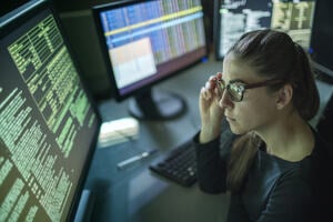 A young woman is seated at a desk surrounded by monitors displaying data