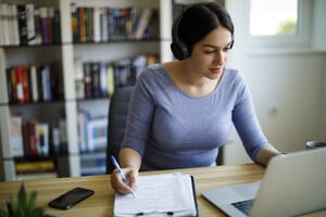 Woman sat at a laptop, while holding a pen over a notepad, wearing headphones