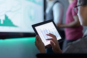 Someone working on a tablet with charts onscreen. A larger screen in the background with two people standing near also shows charts.