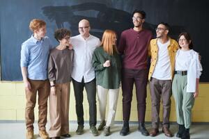 Seven people in business casual dress, a variety of heights, genders, ages and ethnicities stand in a line in front of a chalkboard. They are smiling, and have their arms around each other's shoulders and backs.